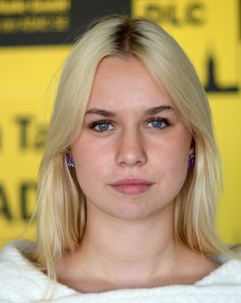 Junge Frau mit blonden Haaren schaut ernst in die Kamera vor gelbem Hintergrund