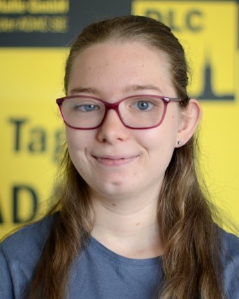 Junge Frau mit langen braunen Haaren und Brille lächelt vor gelbem Hintergrund