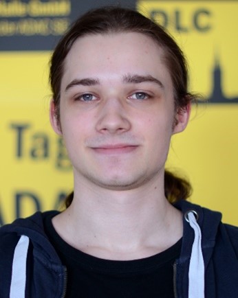 Junger Mann mit langen braunen Haaren und freundlichem Gesichtsausdruck vor gelber Wand